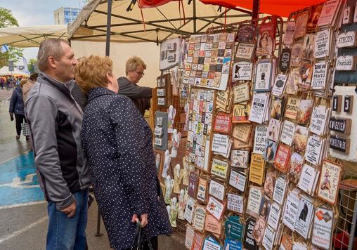 Stoisko z ozodobnymi plakietakami wywieszonymi na dużej płycie. Ogląda je kobieta z mężczyzną