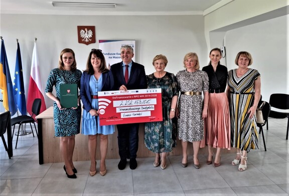 Elżbieta Lanc i Janina Ewa Orzełowska pozują wraz z przedstawicielami gminy Sarnaki do zdjęcia z czekiem. Czek trzyma stojący w środku mężczyzna