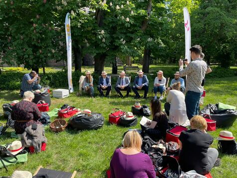 Dziennikarze siedzą na łące i notują, mężczyźni w słomkowych kapeluszach siedzą na leżakach