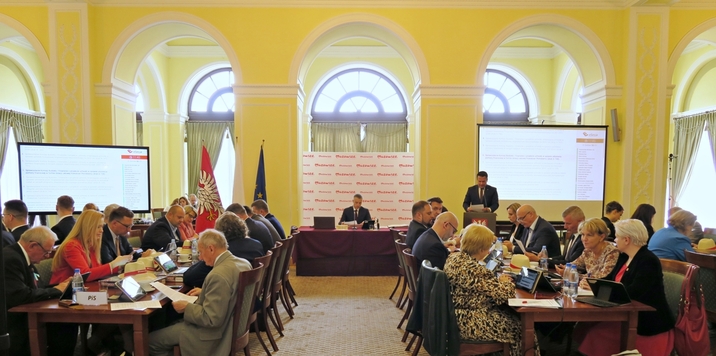 Sala obrad Sejmiku Województwa Mazowieckiego. Po bokach radni siedzą przy stołach, na wprost przy stole prezydialnym siedzi przewodniczący Ludwik Rakowski. Obok stołu prezydialnego, przy mównicy stoi Mirosław Adam Orliński.