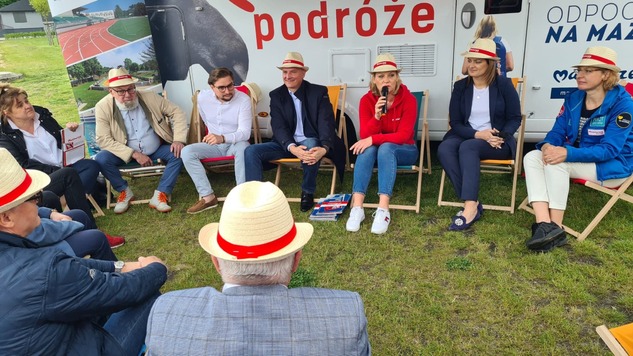 Grupa osób w słomkowych kapeluszach siedzi na leżakach. W tle kamper z logo Mazowsza