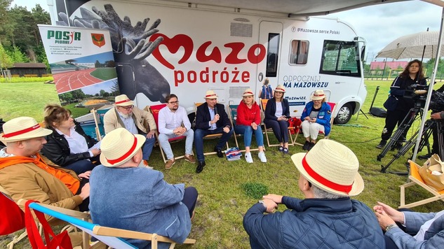 Grupa osób w słomkowych kapeluszach siedzi na leżakach. W tle kamper z logo Mazowsza