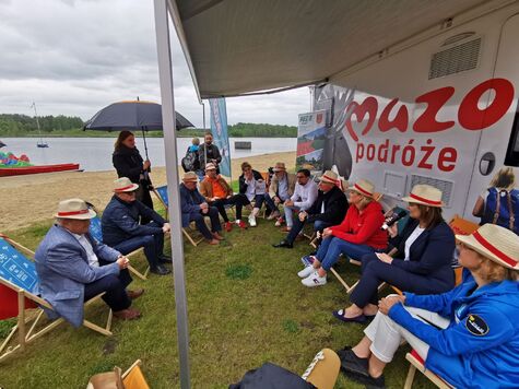 Grupa osób w słomkowych kapeluszach siedzi na leżakach. W tle kamper z logo Mazowsza