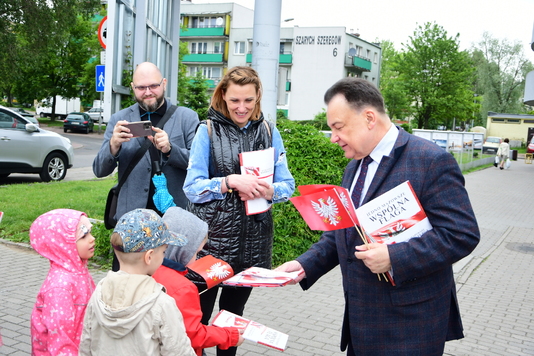 Mężczyzna w garniturze rozdaje chorągiewki z flagą Mazowsza dzieciom