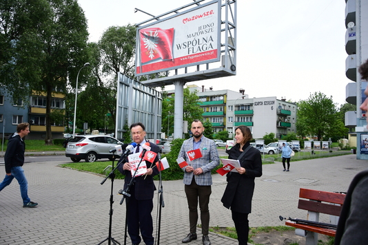 Konferencja prasowa - mężczyźni mówią do stojących przed nim mikrofonów. W tle bilboard z Flagą Mazowsza.