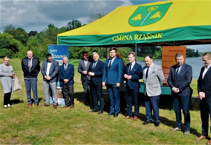 Na łące, nad brzegiem rzeki pod namiotem z napisem gmina Rząśnik stoi marszałek Struzik, radny Augustyniak oraz przedstawiciele lokalnych władz.