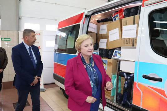 Elegancko ubrana kobieta i mężczyzna w garniturze obok karetki zapakowanej po brzegi w sprzęt medyczny.