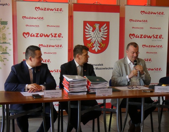 Trzech mężczyzn siedzi przy stole na tle ścianki z logiem Mazowsza i z herbem Mazowsza