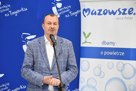 Mężczyzna w garniturze mówi do mikrofonu na tle błękitnych i białych ścianek