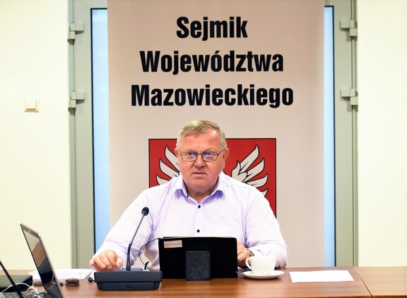 Przewodniczący siedzi za stołem obrad i patrzy przed siebie. Przed nim na stole tablet, mikrofon, laptop, filiżanka, papiery. Za nim roll-up z napisem Sejmik Województwa Mazowieckiego i godłem Mazowsza