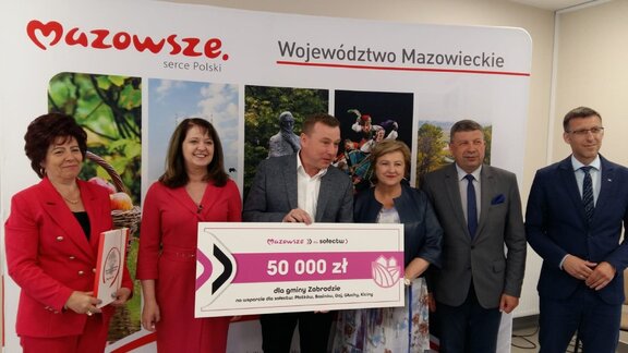 Janina Ewa Orzełowska i Elżbieta Lanc oraz radny Augustyniak pozują do zdjęcia z beneficjentami z gminy Zabrodzie. Jeden z mężczyzn trzyma symboliczny czek na 50 tys. zł