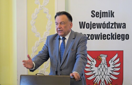 Marszałek przemawia do mikrofonu z trybuny sejmiku, gestykuluje prawą ręką, ręka lewa oparta o pulpit. Za nim roll-up z godłem Mazowsza i napisem Sejmik Województwa Mazowieckiego.