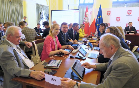 W dużym planie: kilkunastu radnych siedzi z obu stron dwóch stołów podczas obrad. W głębi za nimi ekran dużego telewizora, flagi Polski, Mazowsza i Unii Europejskiej, ścianka z logo Mazowsza i napisami Sejmik Województwa Mazowieckiego.