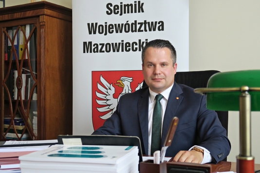 Wiceprzewodniczący sejmiku siedzi za biurkiem. Przed nim mikrofon, teczka i dokumenty, tablet., lampa. Za nim roll-up z herbem Mazowsza i napisem Sejmik Województwa Mazowieckiego oraz szafa oszklona z dokumentami.