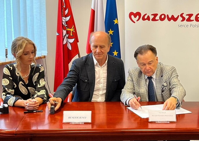 Marszałek siedzi przy stole i podpisuje dokumenty, które leżą przed nim. Obok siedzi mężczyzna i kobieta. W tle są flagi Mazowsza, Polski oraz Unii Europejskiej