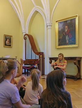 Przed grupą dzieci siedzących na podłodze, siedzi na krześle dziewczyna z gitarą. Zajęcia odbywają się w muzealnej sali