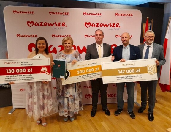 Elżbieta Lanc pozuje do zdjęcia z trzema mężczyznami w garniturach i jedną kobietą - beneficjentami, z którymi podpisywała jedną z umów. Mężczyźni i kobieta trzymają trzy symboliczne czeki, a Elżbieta Lanc dodatkowo trzyma teczkę aktową