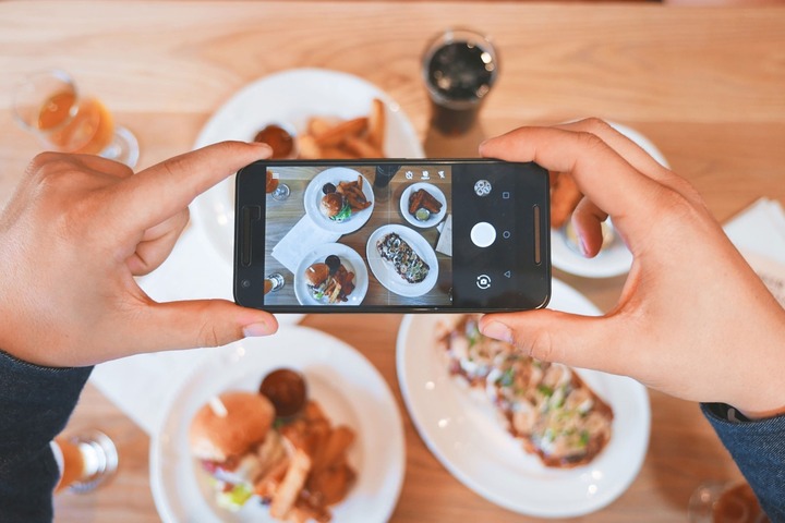 Dwie dłonie trzymają telefon komórkowy. Na jego ekranie wyświetla się fotografowane z góry danie rozłożone pod spodem na czterech talerzach