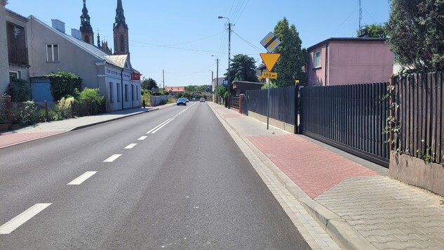 Wyremontowana droga, obok widoczne znaki drogowe i budynki.