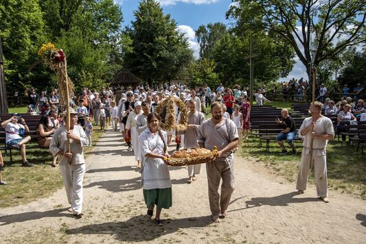 Na pierwszym planie kobieta i mężczyzna w wiejskich strojach niosą wielki bochen chleba, za nimi w korowodzie ludzie niosą wieniec ze ściętego zboża