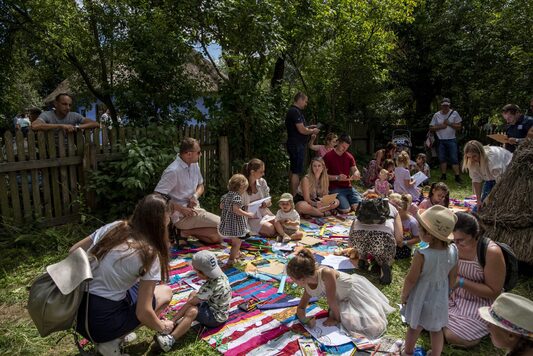 Dzieci siedzą na dywanikach rozłozonych na trawie. Pochylają się nad wykonywanami przez siebie maskami