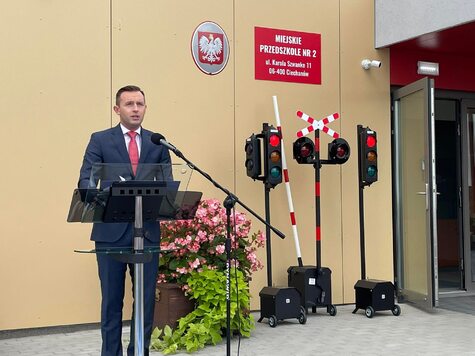 Elegancko ubrany mężczyzna przemawia przez mikrofon na tle wejścia do przedszkola