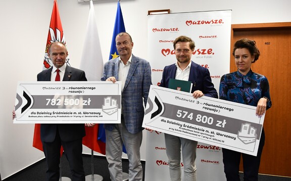 Wicemarszałek Raboszuk stoi wraz z trzema przedstawicielami beneficjenta i pozuje do zdjęcia. Trzymają przed sobą dwa symboliczne czeki