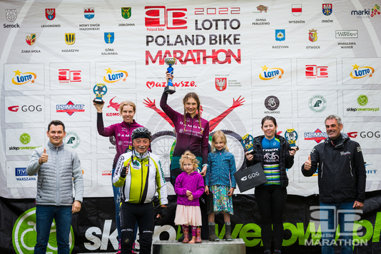 Osiem osób pozuje z na ściance Lotto Poland Bike Marathon. Jedna kobieta trzyma w ręku puchar, a dwie statuetki Lotto. Wśród zebranych radny województwa Krzysztof Skolimowski.