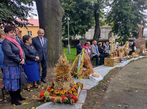 Na placu stoją w rzędzie wieńce dożynkowe. Za nimi stoją osoby, które je wykonywały.