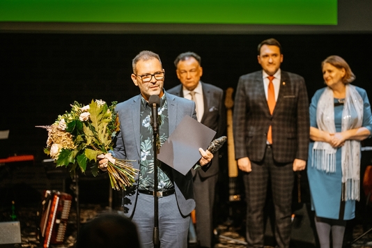 Mariusz Godlewski stoi przy mikrofonie. W prawym ręku trzyma kwiaty, w lewym ręku dyplom i statuetkę. W tle widać marszałka Adama Struzika, wiceprzewodniczącego Marcina Podsędka i przewodniczącą Komisji Kultury i Dziedzictwa Narodowego Jadwigę Zakrzewską.