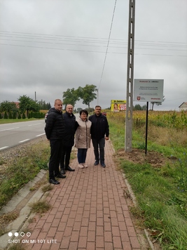 Grupa osób stojąca na chodniku i pozująca do zdjęcia przy tablicy informującej o projekcie. 