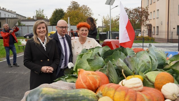 Trzy osoby pozują przy przyczepie z warzywami. W tle mężczyzna robi zdjęcie telefonem komórkowym kobiecie.