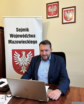 Przy biurku, przed laptopem siedzi wiceprzewodniczący Marcin Podsędek. Za nim stoi roll-up z napisem Sejmik Województwa Mazowieckiego i herbem Mazowsza. Na ścianie w ramkach wiszą godło Polski i herb Mazowsza.