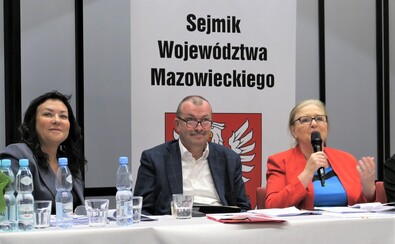 Wicemarszałek Raboszuk siedzi za stołem prezydialnym na posiedzeniu sejmiku. Za nim jest baner z godłem Mazowsza i pełną nazwą sejmiku