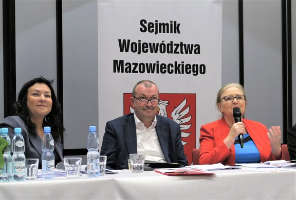 Wicemarszałek Raboszuk siedzi za stołem prezydialnym na posiedzeniu sejmiku. Za nim jest baner z godłem Mazowsza i pełną nazwą sejmiku