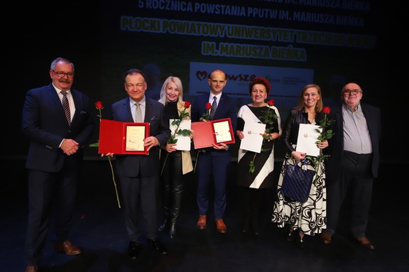 Marszałek Adam Struzik pozuje na scenie do zdjęcia razem z innymi jubilatami. Wszyscy trzymają dyplomy i róże