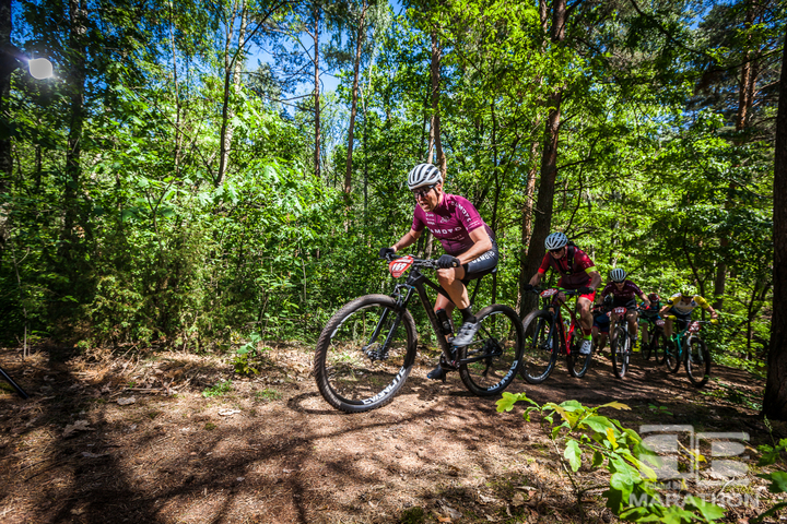 Zawodnik na rowerze mtb jedzie leśną ścieżką pod górę. Za nim widać innych zawodników