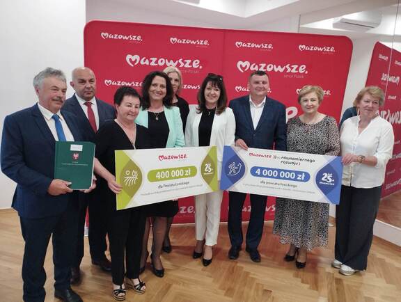 9 osób stoi na tle czerwonej ściany z napisem Mazowsze i prezentują dwa duże czeki z kwotami