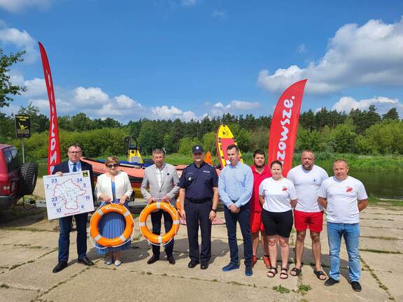 Przedstawiciele samorządu Mazowsza, WOPR, policji stoją na tle wody i prezentują mapę Mazowsza z zaznaczonymi czarnymi punktami wodnymi