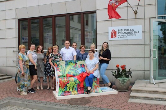 dyrektor Krusiewicz i pracowniczki biblioteki rozwiązują kokardę związana na ławeczce