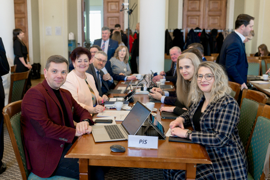 Grupa uśmiechniętych radnych w oczekiwaniu na rozpoczęcie obrad.