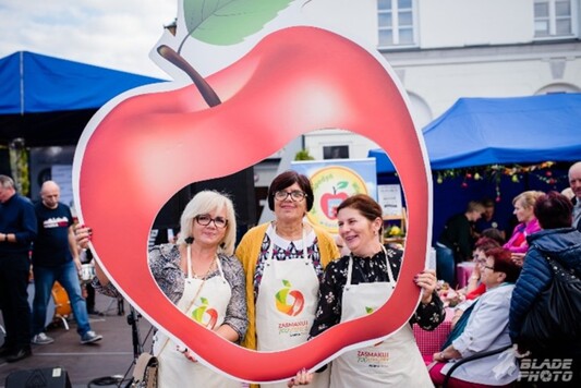 Trzy uśmiechnięte kobiety z planszą przedstawiającą olbrzymie jabłko.