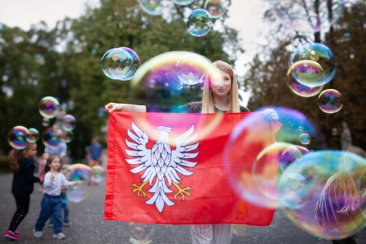 Młoda kobieta trzymająca flagę Mazowsza, stojąca wśród dzieci radośnie łapiących bańki mydlane.