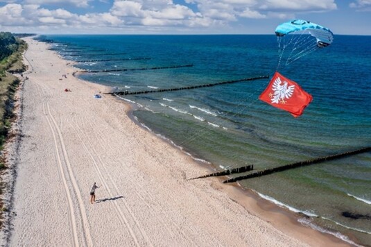 Mężczyzna stojący na nadmorskiej plaży, trzymający wysoko nad nim uniesiony latawiec z dołączoną do niego flagą Mazowsza.