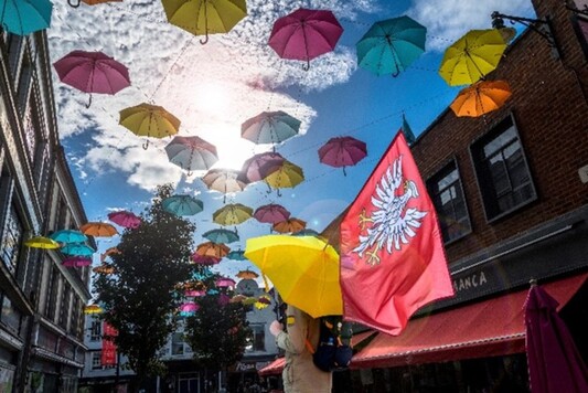 Flaga Mazowsza sfotografowana na tle nieba i kolorowych parasolek.