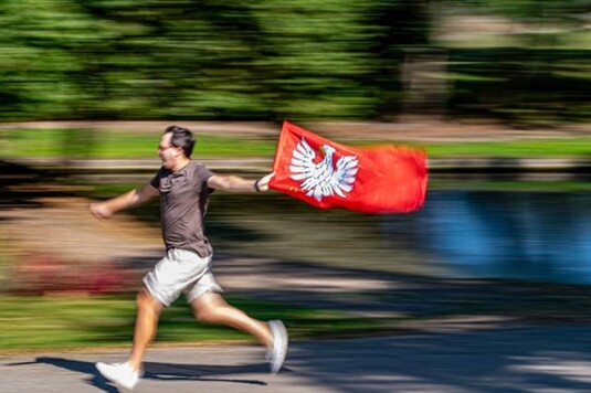 Mężczyzna biegnący z flagą Mazowsza. Na drugim planie jest widoczny rozmyty las, sprawiający wrażenie bardzo szybkiego ruchu mężczyzny z pierwszego planu.