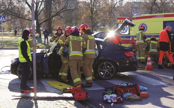 Pracownicy Państwowej Straży Pożarnej przy samochodzie, który uległ wypadkowi. W tle stojący ambulans pogotowia ratunkowego.