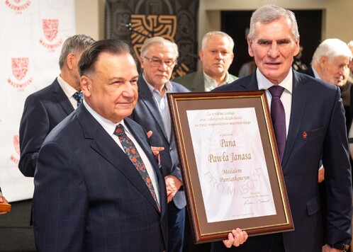 Dwaj mężczyźni w garniturach stoją razem. Jeden z nich trzyma duży dyplom w ramce. W tle widać kilka innych osób.