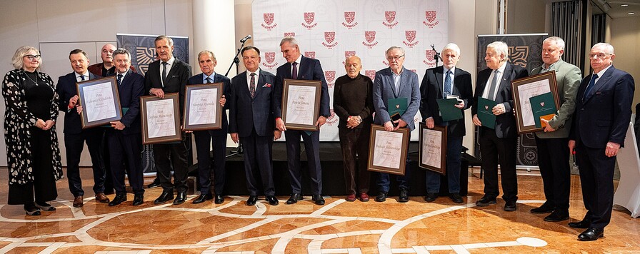 Grupa kilkunastu osób stoi obok siebie, niektóre trzymają duże, oprawione dyplomy i teczki. W tle widoczne są bannery z powtarzającymi się emblematami. Podłoga ma geometryczny wzór.