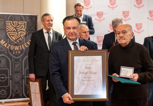 Grupa mężczyzn w garniturach podczas ceremonii wręczenia nagród. Jeden z nich trzyma duży certyfikat w ramie, a inny medal w eleganckim etui. W tle banery z napisami.